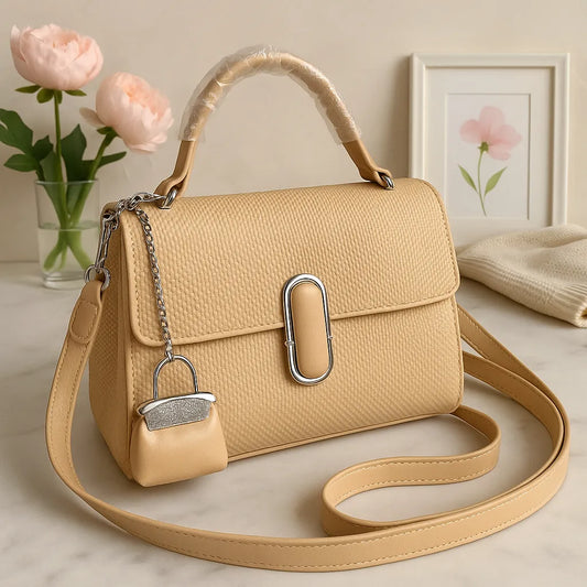Beige handbag with a matching strap on a light surface, with flowers and a framed picture in the background.
