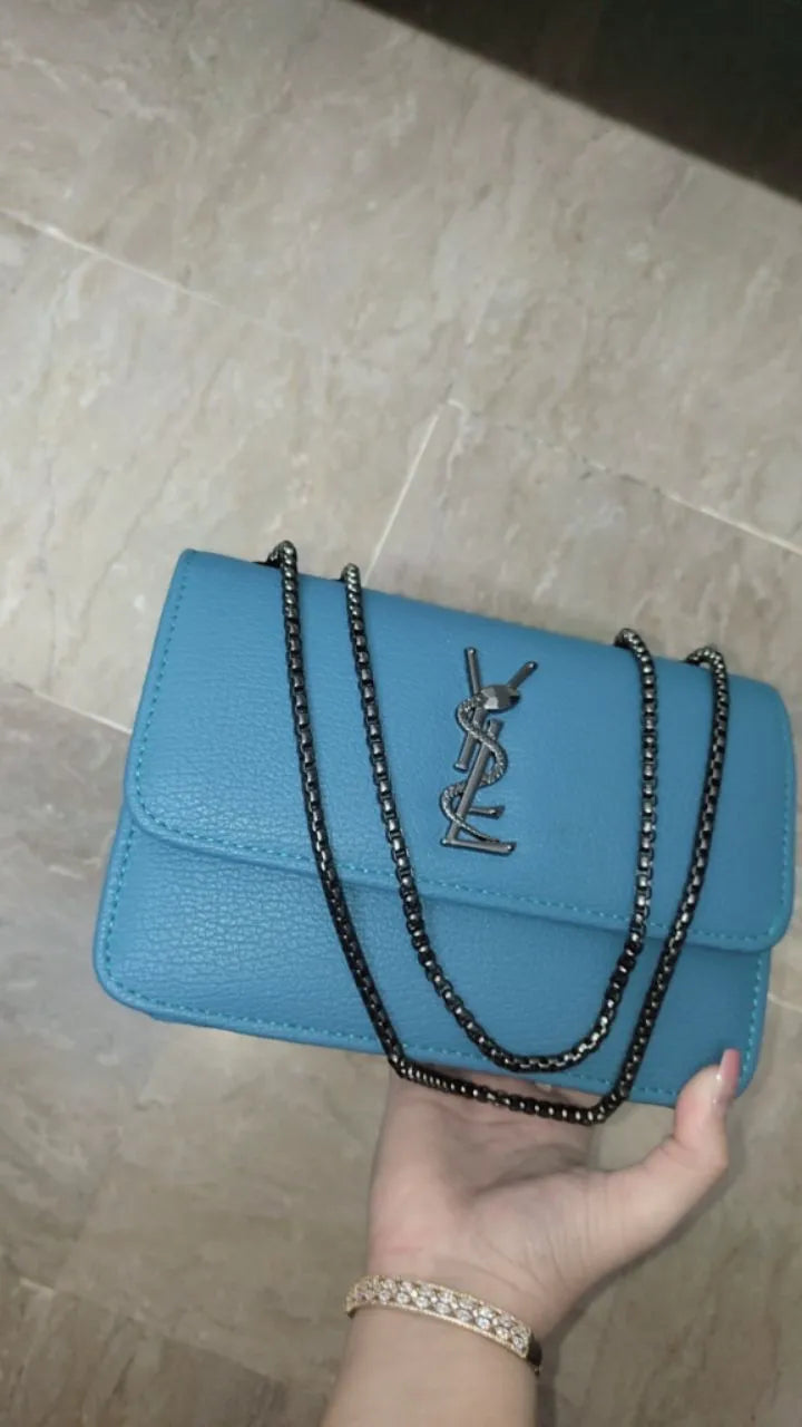 Blue handbag with chain strap held by a hand on a tiled floor.
