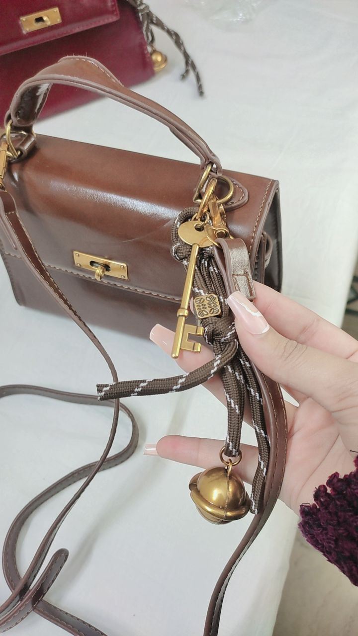 Brown handbag with gold keychain and bell held by a hand on a light background