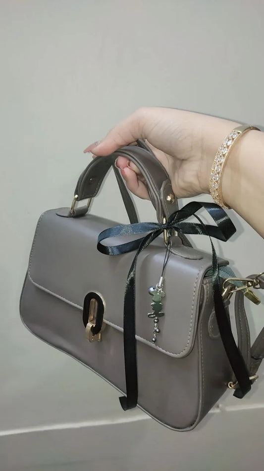 Gray handbag with a black ribbon held by a hand against a neutral background