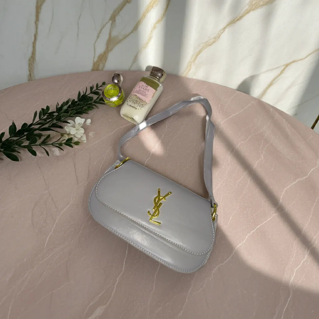 Light gray handbag with a logo on a marble surface with flowers and bottles.